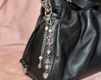 May include: A black leather handbag with a silver chain and a decorative charm. The charm features pink and clear crystal accents, pearls, and a crescent moon. The bag is on a pink surface.