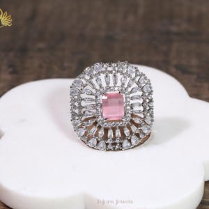 May include: A silver-toned ring with a large, square, light pink center stone. The ring is surrounded by smaller, clear stones in a halo design. The ring is set on a white, flower-shaped surface. The brand name "Tejora Jewels" is visible.