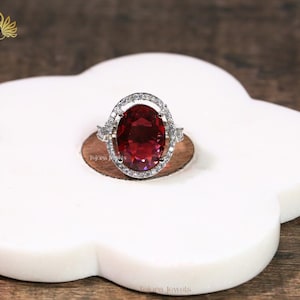 May include: A silver ring with a large, oval-cut, deep red gemstone. The gemstone is surrounded by a halo of smaller, clear stones. The ring is set on a white, cloud-shaped surface. The brand name "Tajora Jewels" is visible.