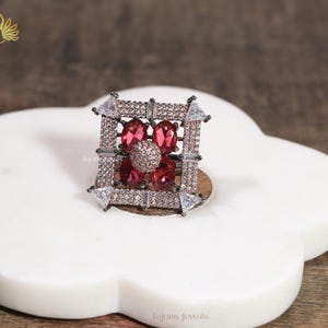 May include: A square-shaped ring with a central cluster of red gemstones surrounded by smaller stones and a square frame. The ring features a mix of clear and red stones, creating a sparkling effect. The ring is set on a white, flower-shaped surface.