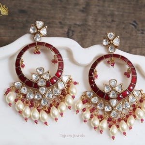 May include: A pair of gold-toned earrings with a circular design. The earrings feature a deep red enamel ring, clear crystal accents, and a cluster of white teardrop-shaped pearls. Small red beads and a floral design complete the look.
