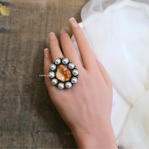May include: A large, ornate ring featuring a central, orange-toned stone surrounded by a halo of small, white pearls. The ring has a silver-colored metal setting. The ring is displayed on a hand.