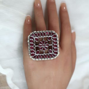 May include: A silver-toned ring with a square design, featuring a central floral motif and surrounding teardrop-shaped red gemstones. The ring is adorned with small, white beads around the outer edge. The ring is a statement piece of jewelry.