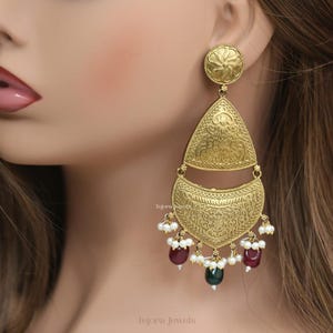 May include: Gold-toned chandelier earrings with intricate embossed patterns. The earrings feature a circular top, a triangular middle section, and a crescent-shaped bottom. They are adorned with white pearls and red and green gemstones.