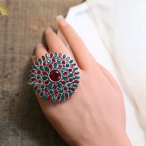 May include: A large, silver-toned ring with a floral design. The ring features a central, round, red gemstone surrounded by alternating red and green gemstones. The ring is displayed on a hand.