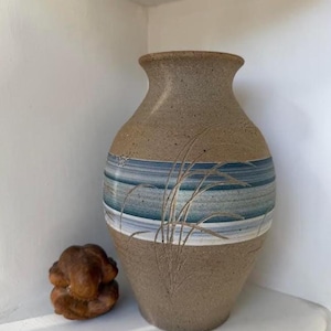 May include: A handcrafted ceramic vase with a wide base and a narrow neck. The vase features a tan body with a band of blue and white horizontal stripes. Delicate etched grass designs are visible on the front. A small, brown, abstract sculpture sits beside the vase.