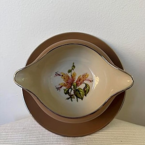 Pode incluir: Uma molheira creme com um desenho floral, colocada sobre um prato a condizer. A molheira tem uma borda prateada e uma pega de cada lado. O prato é de cor castanho claro. O desenho floral apresenta flores rosa e amarelas com folhas verdes.