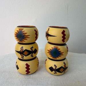 Puede incluir: Dos pilas de tres servilleteros de madera amarillos. Cada anillo está decorado con un diseño diferente de inspiración suroeste en tonos marrones, rojos, azules y verdes. Los anillos están apilados sobre una superficie blanca.