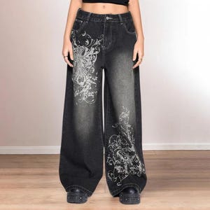 May include: Black wide-leg jeans with a faded wash and white floral patterns. The jeans have a high waist and are worn with black platform shoes. The style is reminiscent of the early 2000s. The jeans are approximately 100 cm in length.