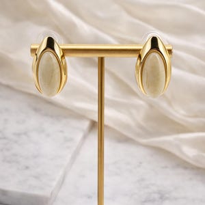 May include: Gold-toned oval stud earrings with a cream-coloured, oval-shaped stone centre. The earrings are displayed on a gold earring stand against a white background.