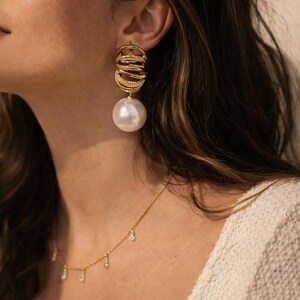 May include: Gold earrings featuring a layered design and a large, white pearl drop. A delicate gold necklace with small, dangling gems is also visible. The jewellery is set against a neutral background.