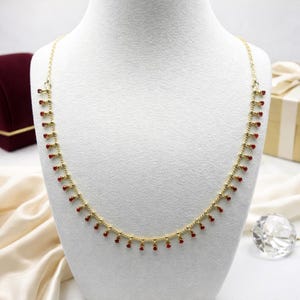 May include: A gold necklace featuring a delicate chain with small, dangling red beads. The necklace is presented on a white mannequin. The background includes a burgundy box, a gift box, and a crystal ornament.