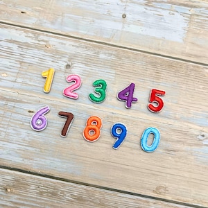 May include: A collection of embroidered number patches in various colors, including yellow, pink, green, purple, red, and orange. Each number is outlined in white thread. The numbers are arranged on a light-colored wooden surface.