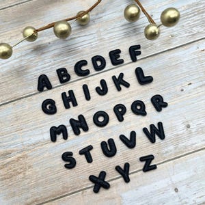 May include: Black felt alphabet letters arranged on a light wood surface. The letters are in a rounded font and are arranged in rows. A gold-colored decorative branch with gold-colored spheres is in the background.