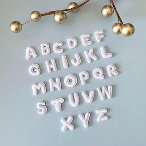 May include: White embroidered alphabet letters arranged on a light blue surface. The letters are in a sans-serif font and are evenly spaced. Decorative gold-colored spheres and a branch are in the background. The letters are approximately 1 inch tall.