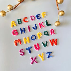 May include: Colorful embroidered alphabet letters in various colors, including red, yellow, blue, and green. The letters are arranged on a light blue surface, with gold decorative accents in the background. The letters are perfect for crafting and personalization.