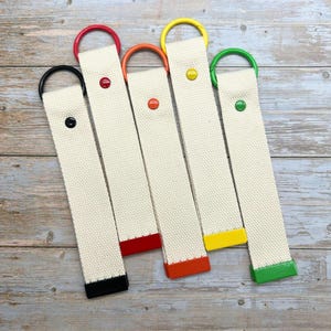 May include: Five cream-colored canvas keychains with colorful accents. Each keychain has a different colored metal ring and end cap: black, red, orange, yellow, and green. A small, matching button secures the canvas.