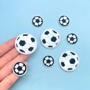 May include: A collection of seven circular embroidered patches featuring a black and white soccer ball pattern. The patches vary in size, with some larger and some smaller, all displaying the classic hexagonal and pentagonal stitching design.