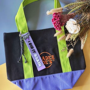 May include: Black tote bag with green handles and accents, and a purple base. The bag features the text "Trader Joe's" in orange embroidery. A purple tag with the text "Oops! I did it again" hangs from the handle.