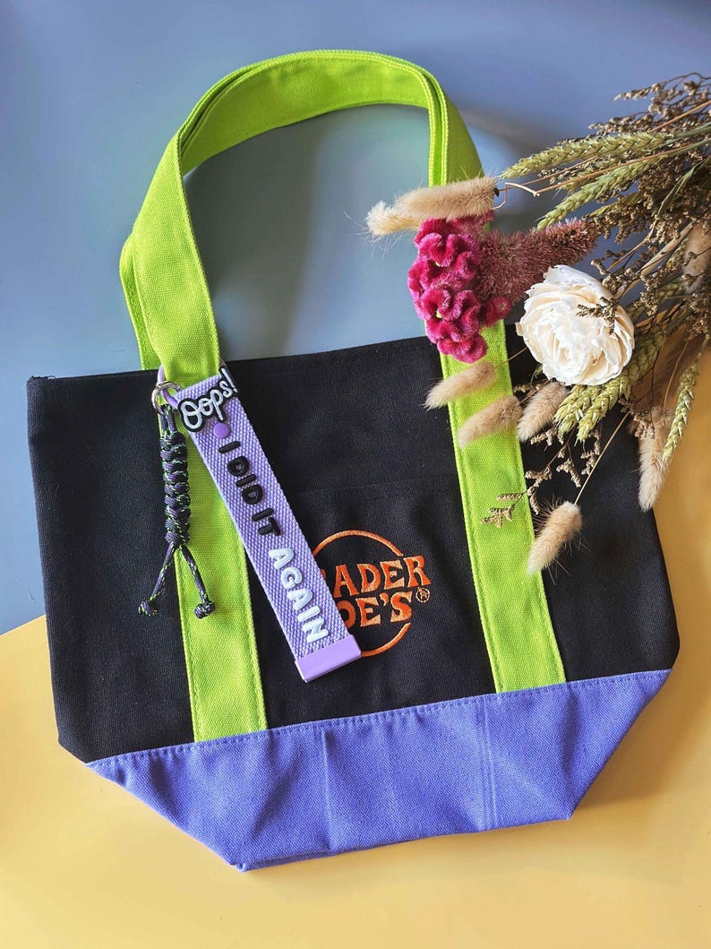 May include: Black tote bag with green handles and accents, and a purple base. The bag features the text "Trader Joe's" in orange embroidery. A purple tag with the text "Oops! I did it again" hangs from the handle.