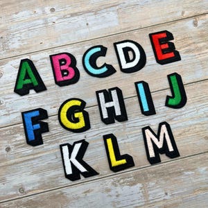 May include: A collection of colorful embroidered alphabet patches. Each letter is outlined in black with a contrasting color fill, including green, pink, blue, white, red, yellow, and light blue. The patches are arranged on a light wood grain surface.