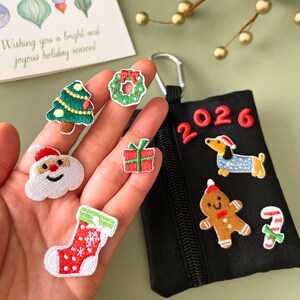 May include: A collection of embroidered Christmas patches, including a Christmas tree, wreath, Santa Claus, gift, stocking, and a black pouch with a dachshund, gingerbread man, candy cane, and the year 2026. A greeting card is in the background.
