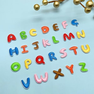 May include: A collection of colorful embroidered alphabet letters, each with a different color and a stitched border. The letters are arranged on a light blue surface, with a few decorative gold orbs in the background. The letters are in a playful, cartoon-like font.