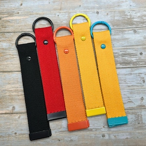 May include: Five colorful canvas keychains in black, red, orange, yellow, and gold. Each keychain has a matching metal ring and a rivet. The keychains are arranged diagonally on a wooden surface.