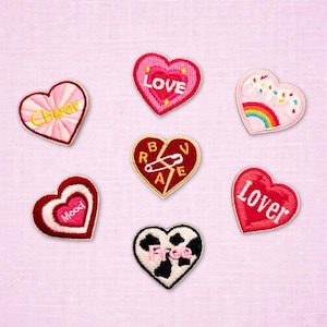 May include: A collection of embroidered heart-shaped patches in various colors. The patches feature words like "Love", "Cheer", "Mood", "Lover", and "Free". Some patches have additional designs, such as a rainbow or a safety pin.