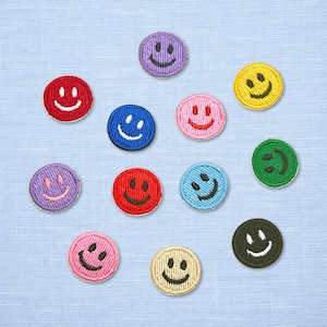 May include: A collection of embroidered smiley face patches in various colors, including red, purple, blue, pink, yellow, green, and tan. Each patch features a stitched smiling face with a white outline, set against a light blue fabric background.