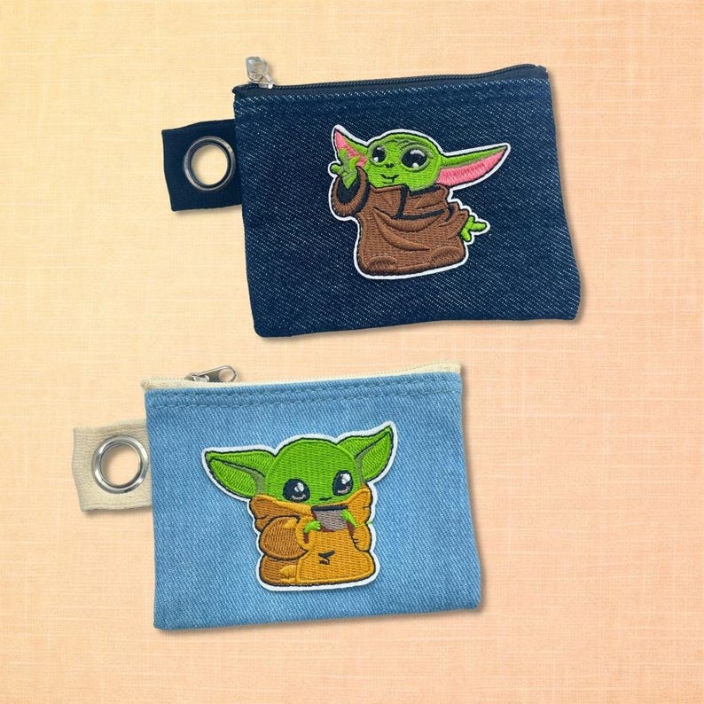 May include: Two denim coin purses with zipper closures and a small loop. Each purse features an embroidered image of a green alien character. One purse is dark blue denim, the other is light blue denim.