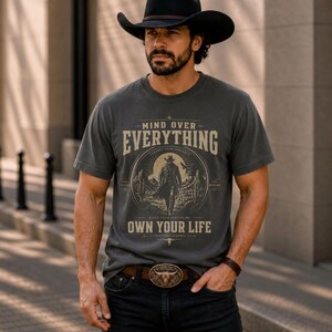 May include: A charcoal gray t-shirt with the text "MIND OVER EVERYTHING" and "OWN YOUR LIFE" in a vintage style. The shirt features a cowboy graphic. The person is wearing a black cowboy hat, a brown belt with a bull buckle, and black jeans.