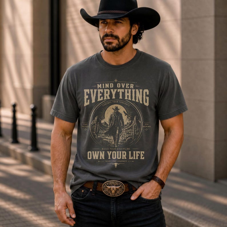 May include: A charcoal gray t-shirt with the text "MIND OVER EVERYTHING" and "OWN YOUR LIFE" in a vintage style. The shirt features a cowboy graphic. The person is wearing a black cowboy hat, a brown belt with a bull buckle, and black jeans.