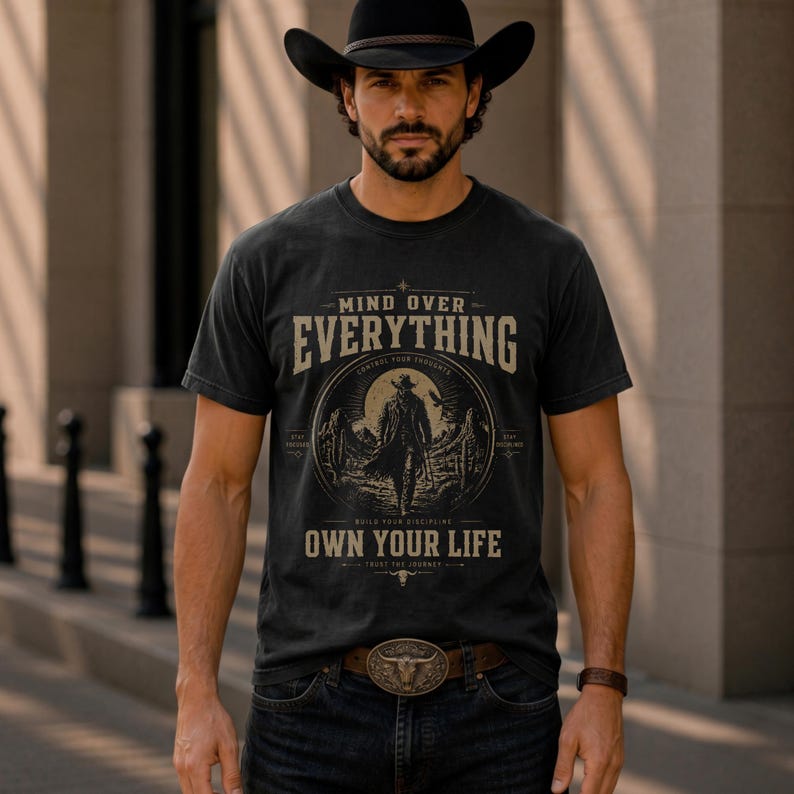 May include: Black t-shirt with the text "MIND OVER EVERYTHING" and "OWN YOUR LIFE". The shirt features a graphic of a cowboy in a desert landscape. The person in the image is wearing a black cowboy hat and a brown belt with a decorative buckle.
