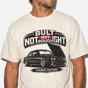 May include: Beige t-shirt featuring a black graphic of a BMW car with the hood open. The text reads "BUILT NOT BOUGHT" above the car and "STANCE CULTURE" below. The design includes red and white accents.