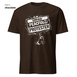Op de afbeelding: Donker chocoladekleurig T-shirt met een witte afbeelding. De afbeelding toont een hand die een bord vasthoudt met de tekst "UNPAID PEACEFUL PROTESTER" en een vredessymbool. Het shirt is een crew neck met korte mouwen.