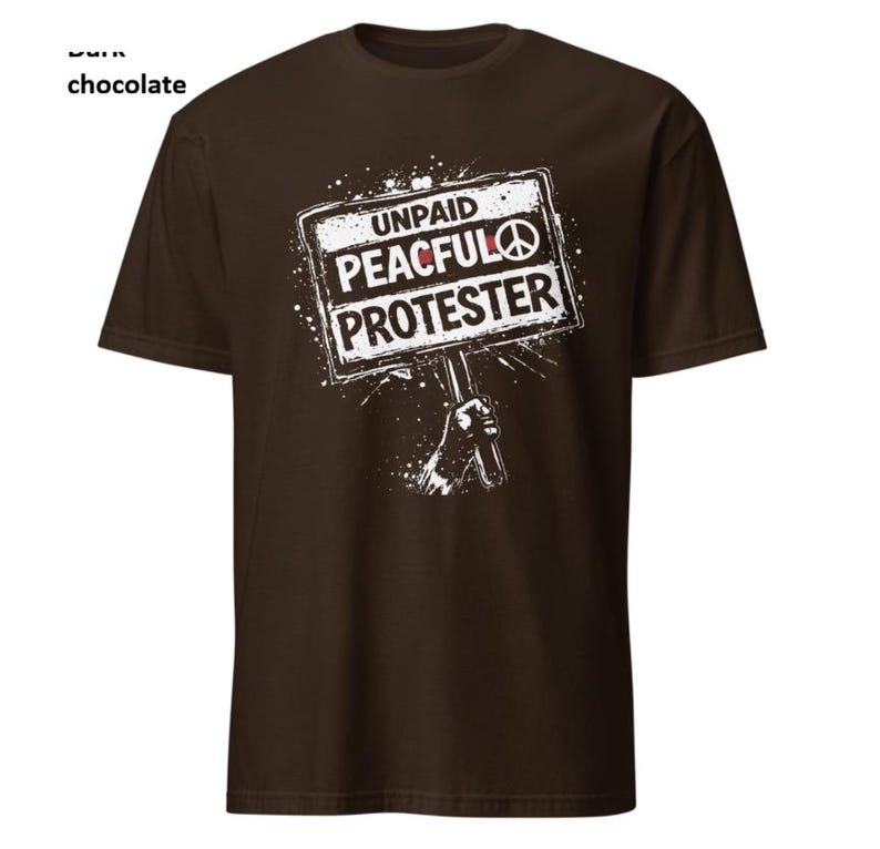 Op de afbeelding: Donker chocoladekleurig T-shirt met een witte afbeelding. De afbeelding toont een hand die een bord vasthoudt met de tekst "UNPAID PEACEFUL PROTESTER" en een vredessymbool. Het shirt is een crew neck met korte mouwen.