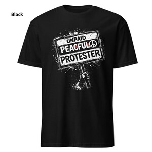Op de afbeelding: Zwart t-shirt met een witte afbeelding van een protestbord. Het bord leest "UNPAID PEACEFUL PROTESTER" met een vredesteken. Het ontwerp bevat een hand die het bord vasthoudt en een spatteffect.
