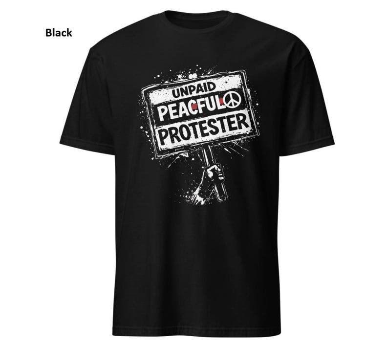 Op de afbeelding: Zwart t-shirt met een witte afbeelding van een protestbord. Het bord leest "UNPAID PEACEFUL PROTESTER" met een vredesteken. Het ontwerp bevat een hand die het bord vasthoudt en een spatteffect.