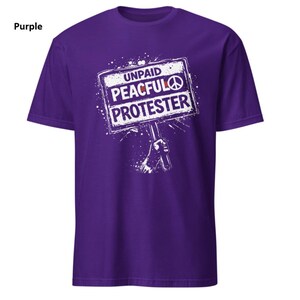 Op de afbeelding: Een paars t-shirt met de witte tekst "UNPAID PEACEFUL PROTESTER" op een bordafbeelding. Het bord bevat een vredessymbool. Het shirt heeft een effen paarse kleur.