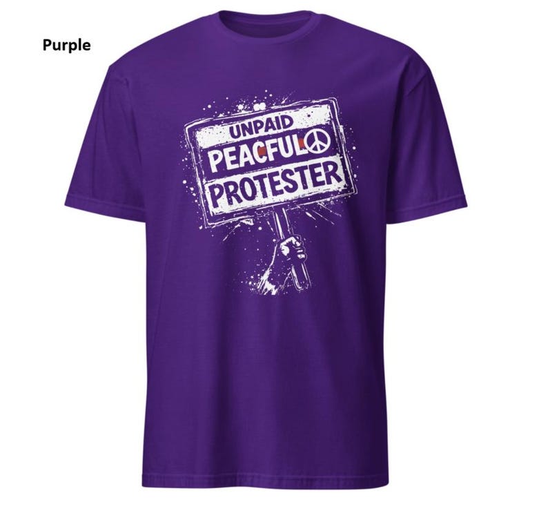 Op de afbeelding: Een paars t-shirt met de witte tekst "UNPAID PEACEFUL PROTESTER" op een bordafbeelding. Het bord bevat een vredessymbool. Het shirt heeft een effen paarse kleur.