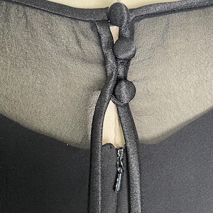 K&ouml;nnte beinhalten: Nahaufnahme eines schwarzen Kleideroberteils mit einem transparenten Overlay. Das Kleid hat drei stoffbezogene Kn&ouml;pfe und einen Rei&szlig;verschluss. Der Ausschnitt ist mit einem satinartigen Material verziert.