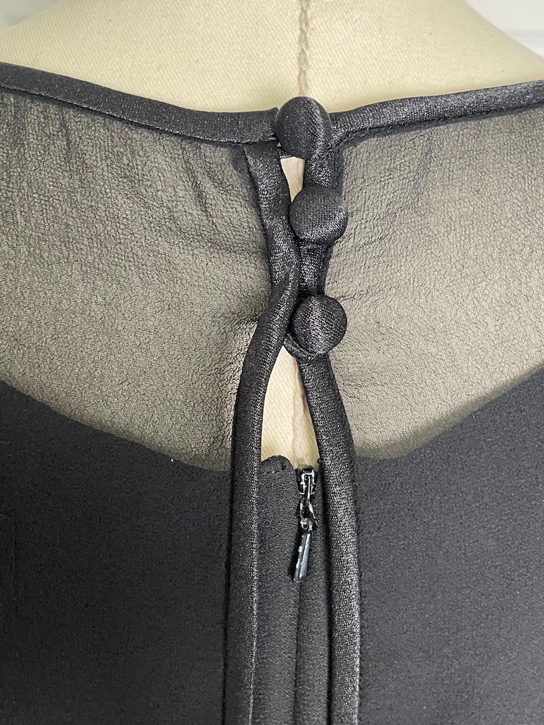 K&ouml;nnte beinhalten: Nahaufnahme eines schwarzen Kleideroberteils mit einem transparenten Overlay. Das Kleid hat drei stoffbezogene Kn&ouml;pfe und einen Rei&szlig;verschluss. Der Ausschnitt ist mit einem satinartigen Material verziert.