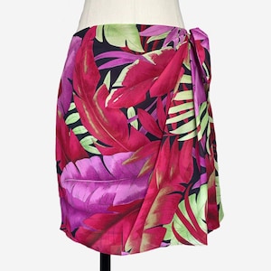May include: A vibrant wrap skirt featuring a tropical leaf print in shades of fuchsia, magenta, and lime green against a black background. The skirt is tied at the side and displayed on a mannequin.