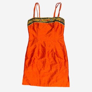 May include: An orange, sleeveless dress with thin straps. The neckline is adorned with a decorative band featuring black, gold, and red embellishments. The dress has a fitted silhouette and is made of a shiny fabric.