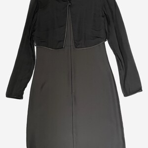K&ouml;nnte beinhalten: Ein dunkelgraues Kleid mit passendem, durchsichtigem, lang&auml;rmligem Bolero. Der Bolero hat einen einzelnen Knopfverschluss am Ausschnitt und eine kontrastierende Paspel. Das Kleid scheint knielang zu sein.