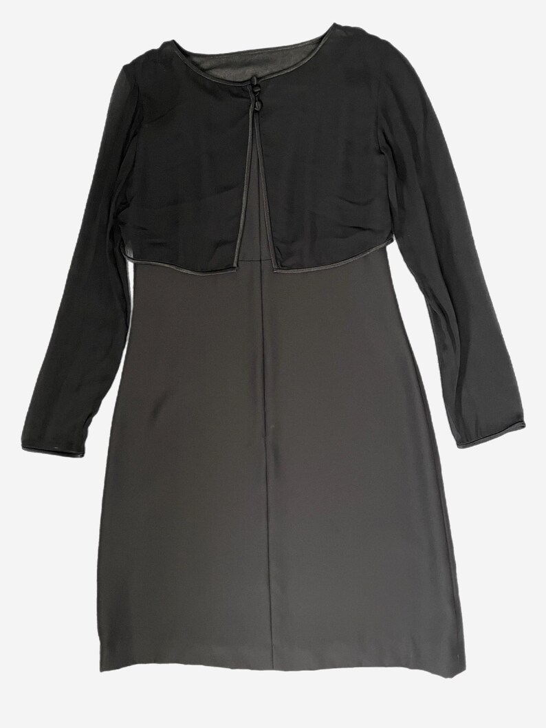 K&ouml;nnte beinhalten: Ein dunkelgraues Kleid mit passendem, durchsichtigem, lang&auml;rmligem Bolero. Der Bolero hat einen einzelnen Knopfverschluss am Ausschnitt und eine kontrastierende Paspel. Das Kleid scheint knielang zu sein.