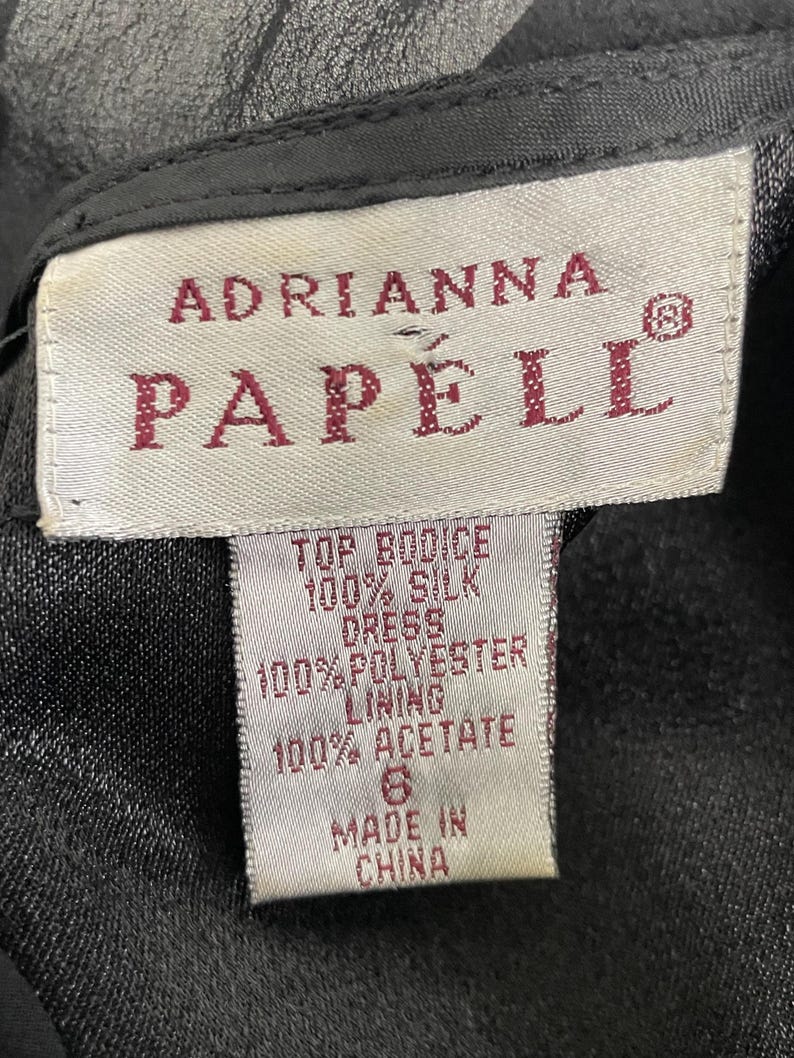 K&ouml;nnte beinhalten: Nahaufnahme eines Etiketts von einem Adrianna Papell Kleid. Das wei&szlig;e Etikett hat burgunderfarbenen Text, der die Zusammensetzung des Kleides auflistet: 100 % Seide f&uuml;r das Oberteil, 100 % Polyester f&uuml;r das Futter und 100 % Acetat. Das Etikett gibt auch die Gr&ouml;&szlig;e 6 und Made in China an.