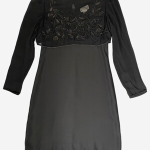 K&ouml;nnte beinhalten: Ein dunkelgraues Kleid mit langen &Auml;rmeln. Die obere Schicht hat ein gesticktes Blumenmuster. Ein Etikett ist in der N&auml;he des Halsausschnitts sichtbar. Das Kleid ist ein schlichtes, elegantes Design.