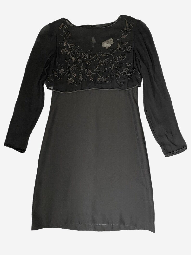 K&ouml;nnte beinhalten: Ein dunkelgraues Kleid mit langen &Auml;rmeln. Die obere Schicht hat ein gesticktes Blumenmuster. Ein Etikett ist in der N&auml;he des Halsausschnitts sichtbar. Das Kleid ist ein schlichtes, elegantes Design.